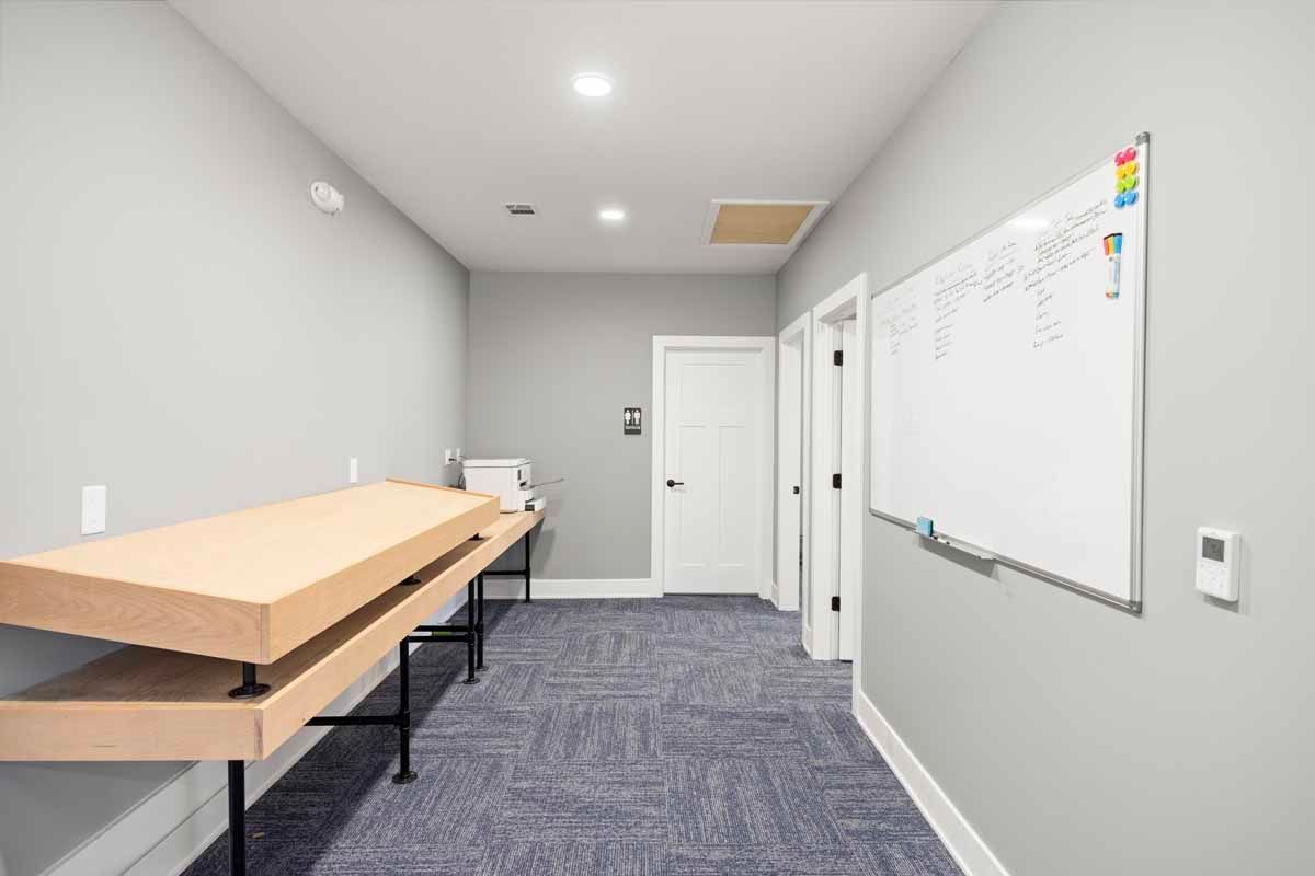 Long, narrow room with light gray walls, carpet, a whiteboard, and a long wooden table.