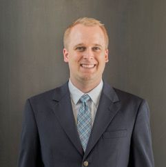 A smiling man in a navy suit and patterned tie against a gray background.
