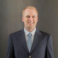 A smiling man with blonde hair wearing a navy suit and blue patterned tie, against a gray background.