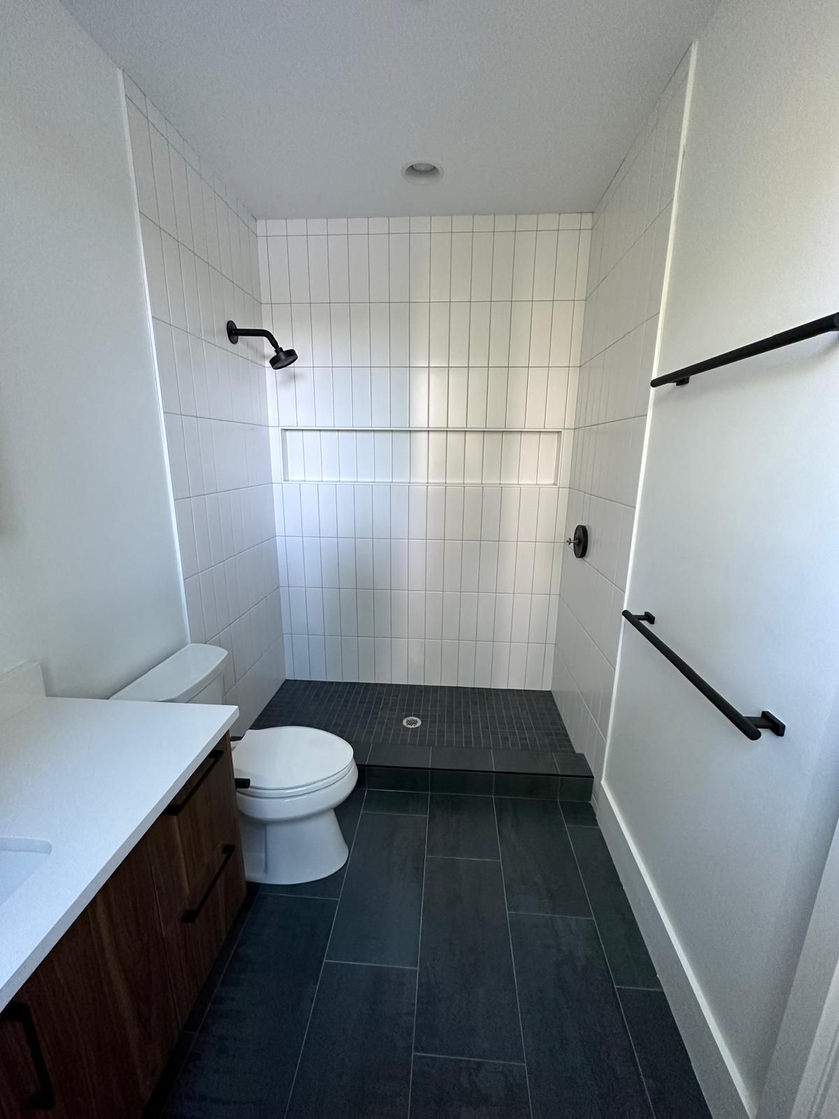 Modern bathroom with white tile shower, dark fixtures, dark gray floors, and a wooden vanity.