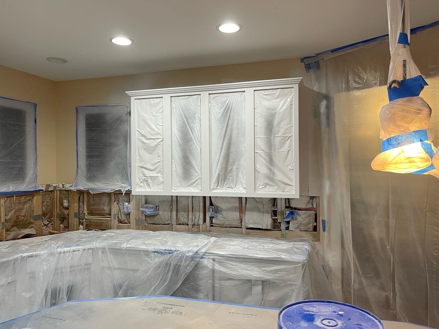 Kitchen renovation: cabinets covered in plastic, walls taped, and a light fixture shielded.