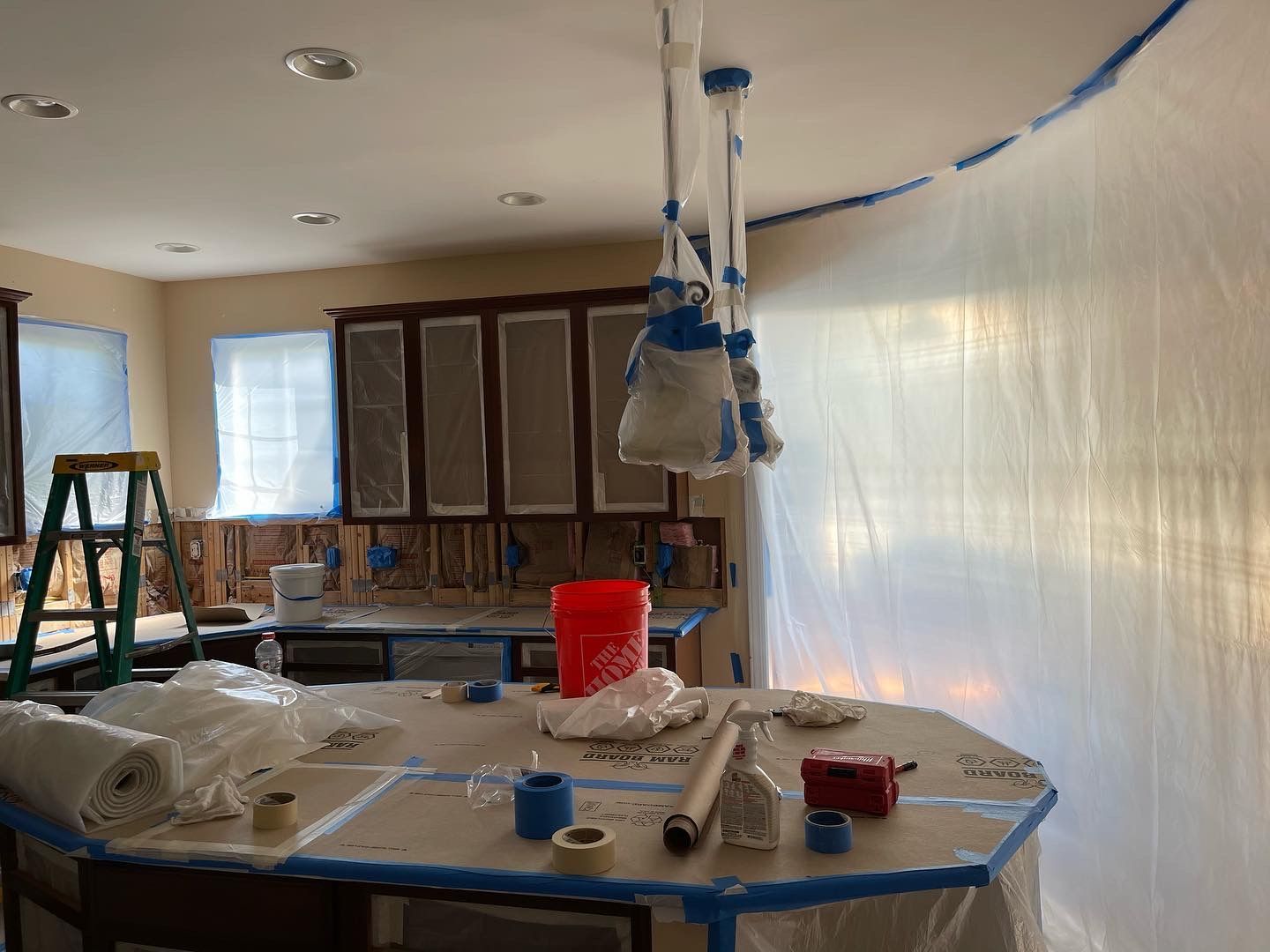 Kitchen under renovation, covered in plastic sheeting and tape. Cabinets, island, and ceiling lights are protected.