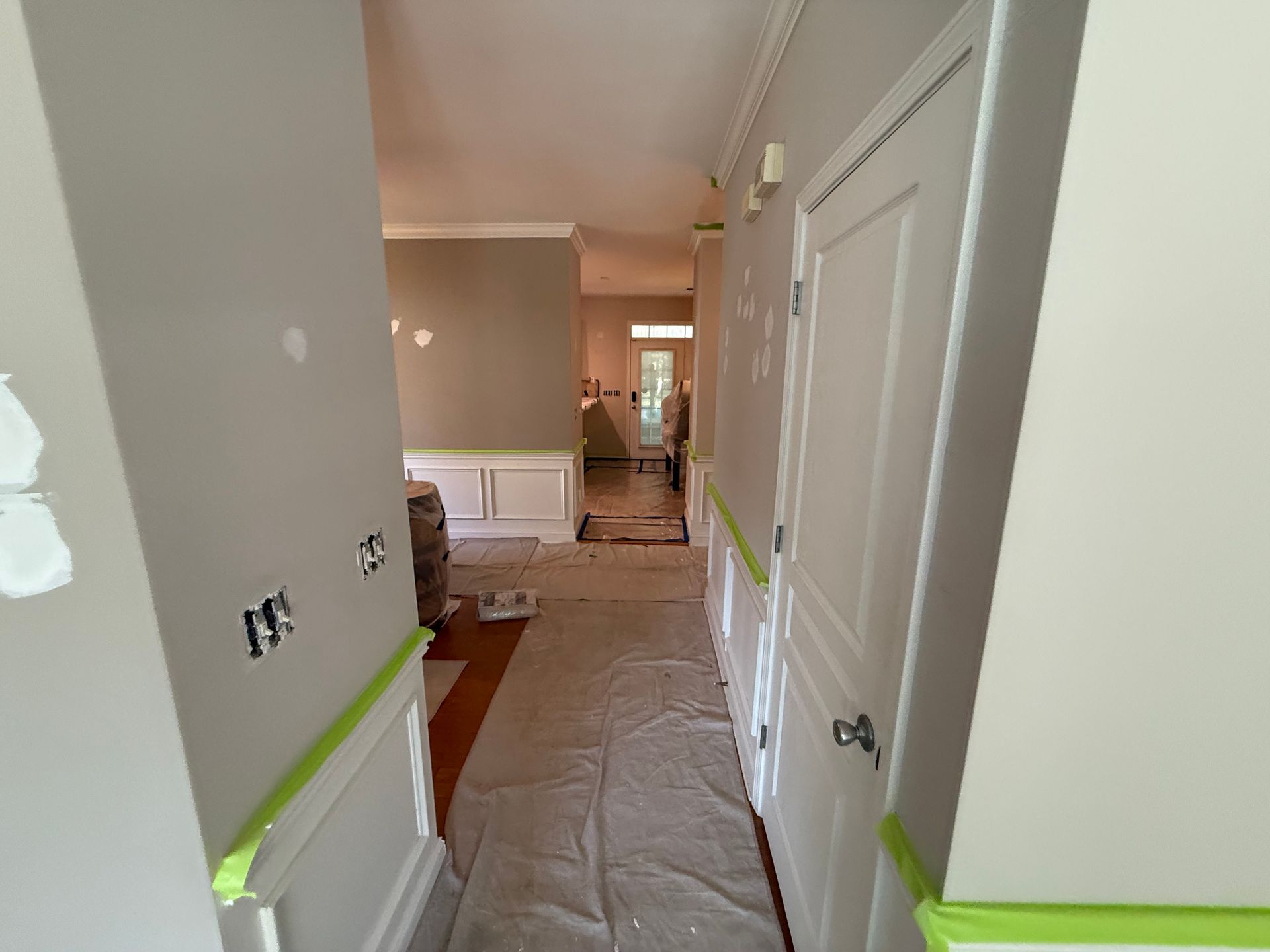 Hallway under renovation, beige walls with white trim, green tape, covered floor, open door at the end.