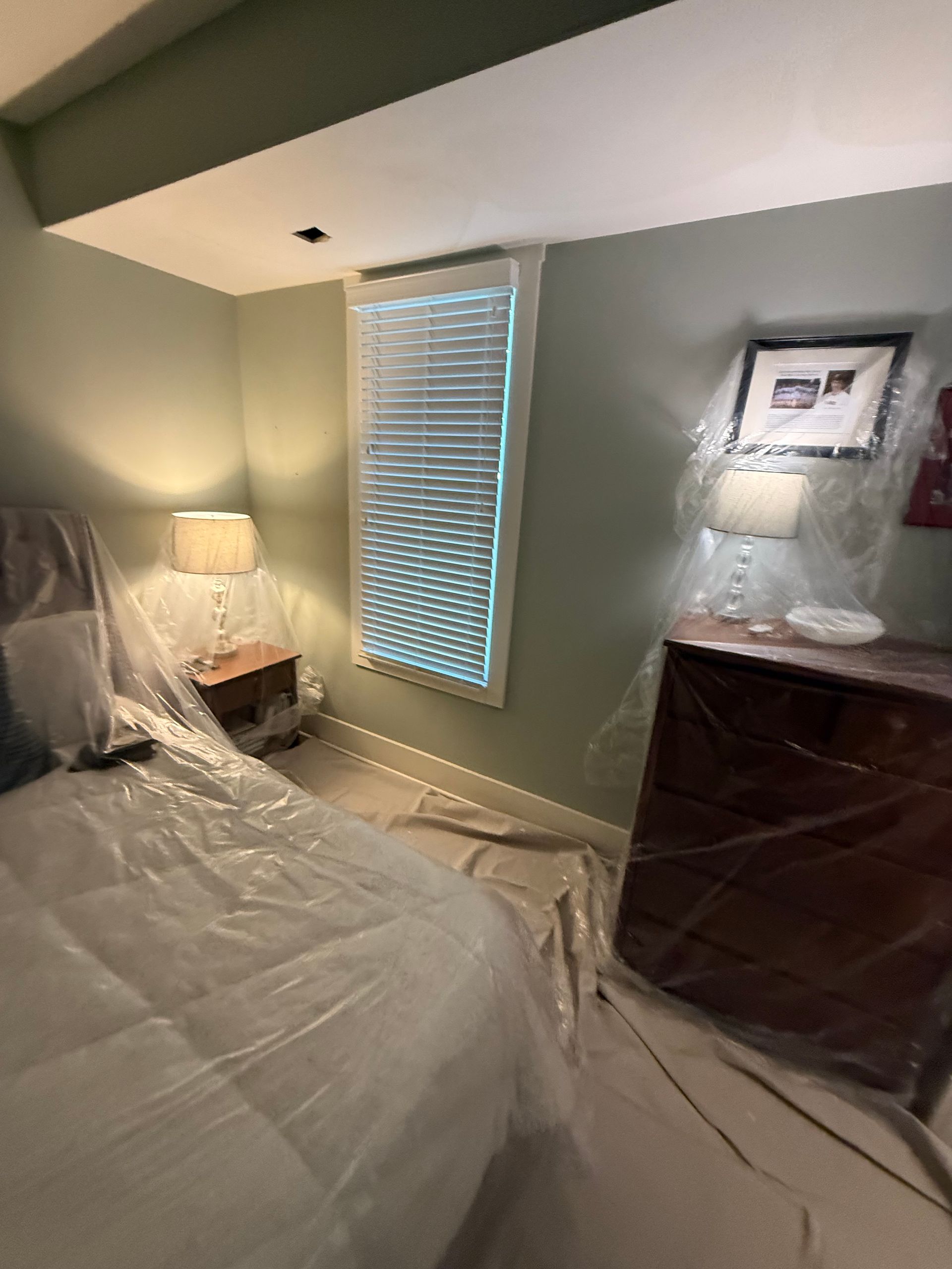 Bedroom with green walls, furniture covered in plastic, window with blinds, lamps on nightstands and dresser.