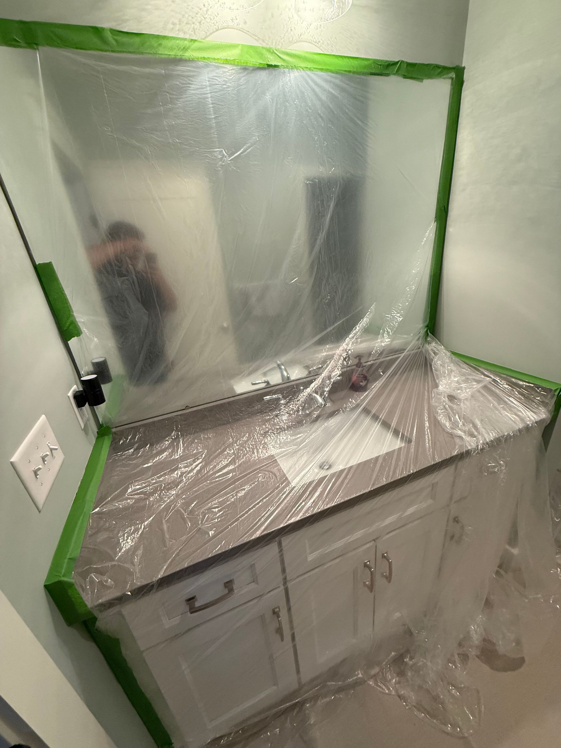 Bathroom vanity covered in plastic sheeting for painting, with a person reflected in the mirror.