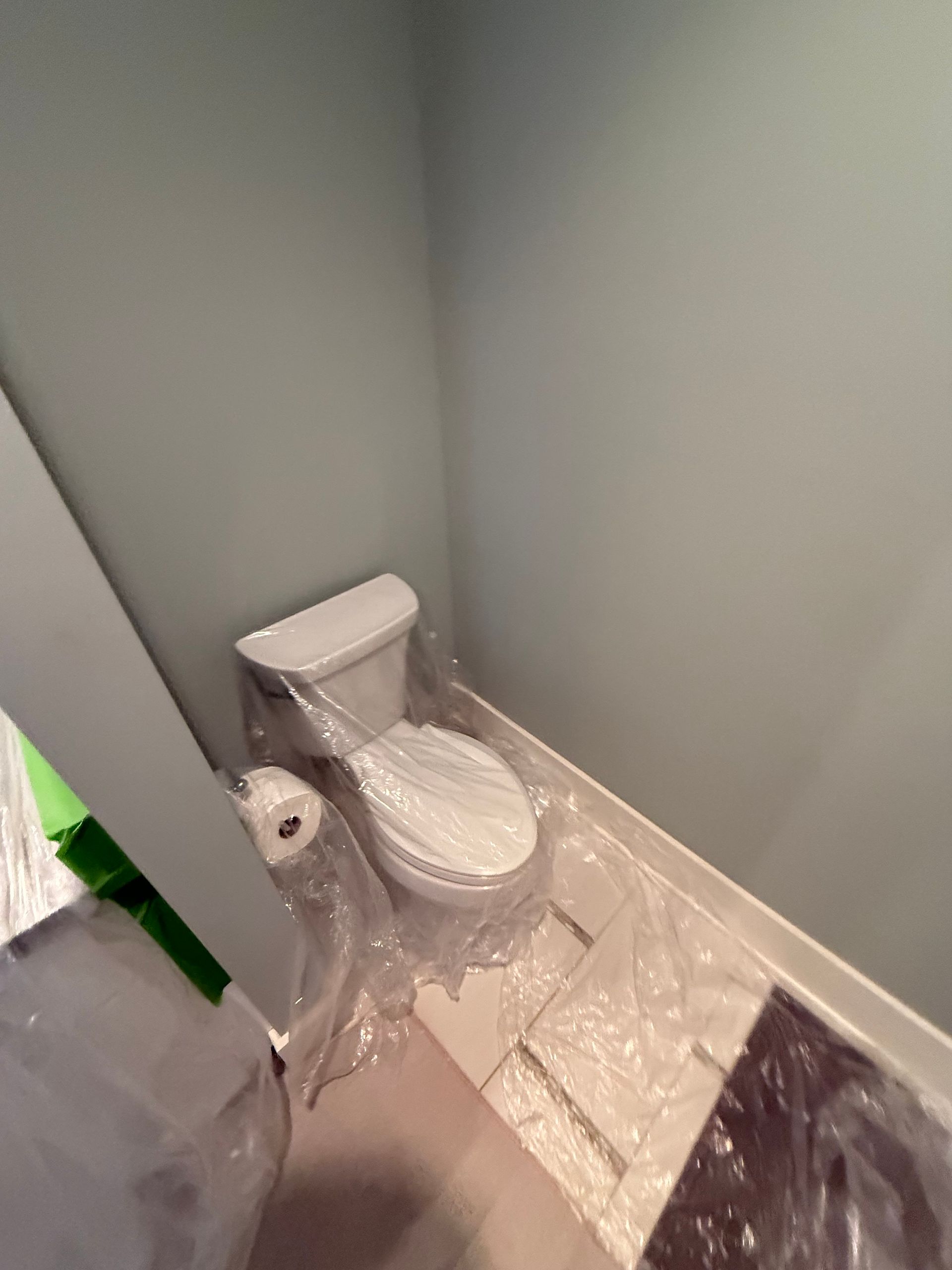 Toilet in a corner of a room, covered in plastic for painting. Gray walls and white trim.