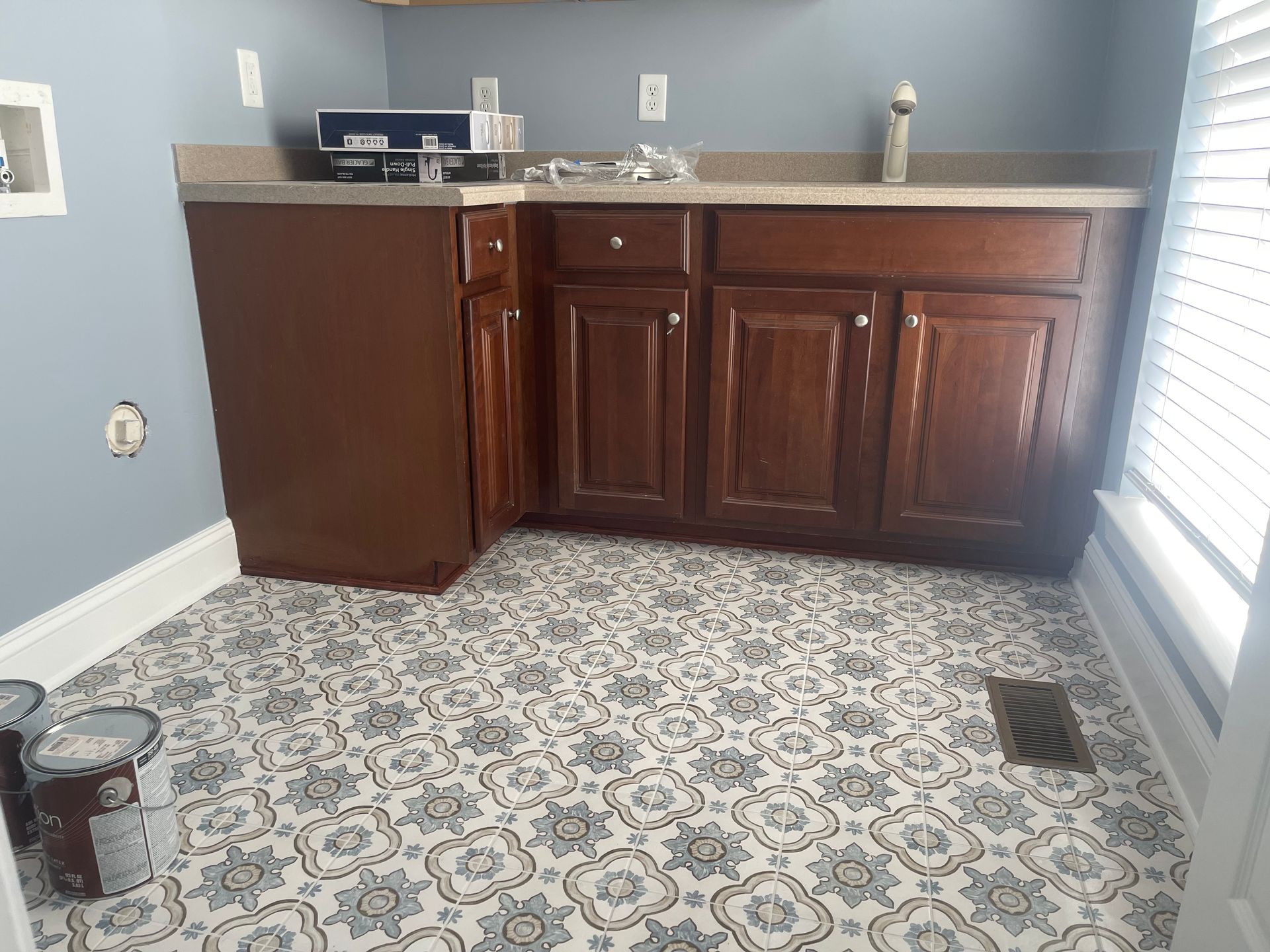 Laundry room with patterned floor and brown cabinets. Blue walls, paint cans, and window.