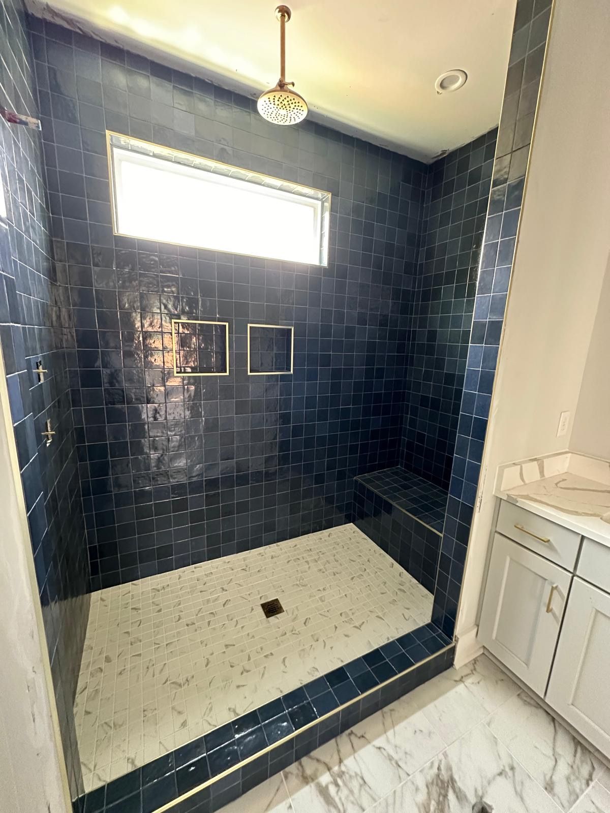 Dark blue tiled shower with white floor and a small window, vanity with a white countertop.