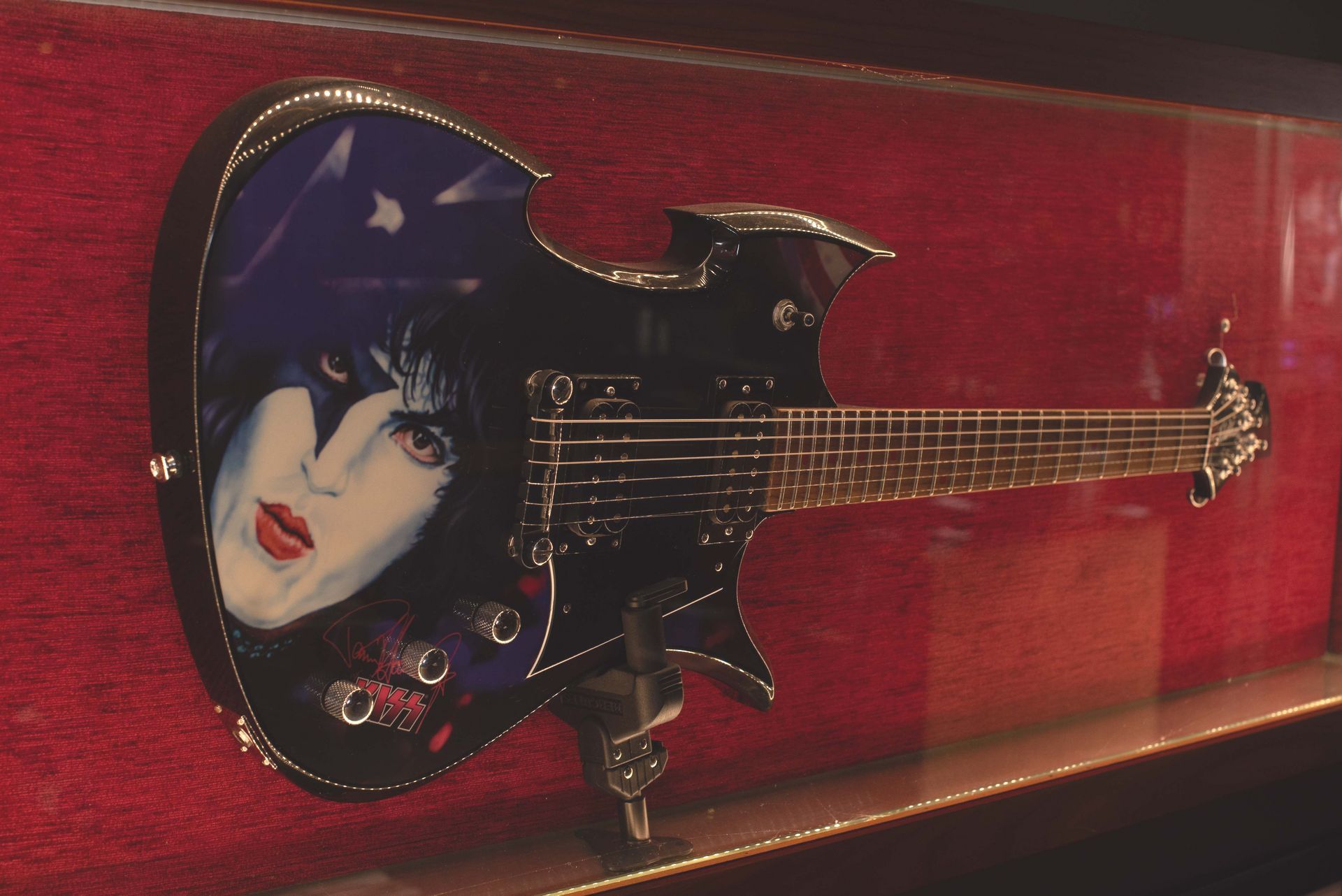 A black electric guitar with a painted face of Gene Simmons, displayed in a red-lined shadow box.