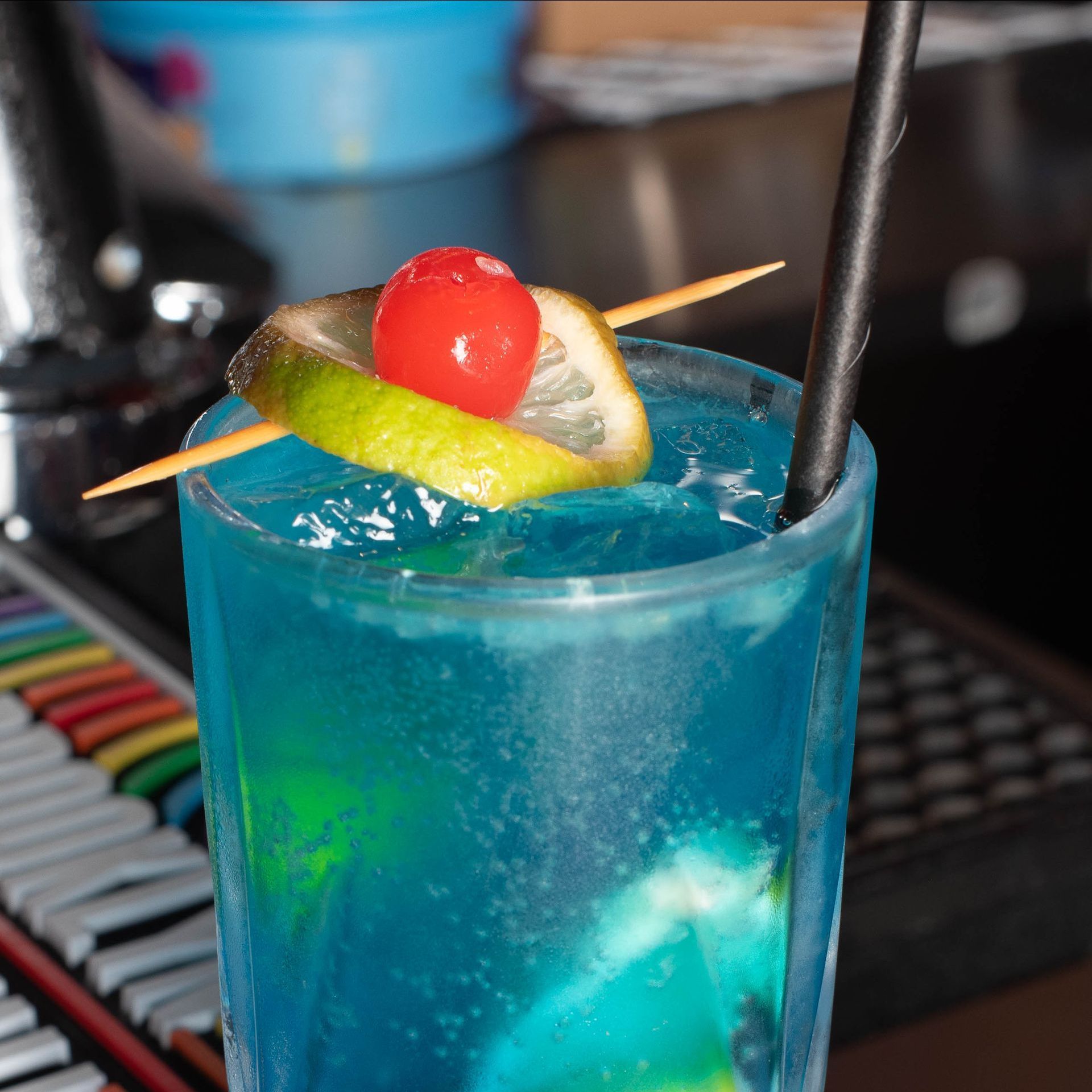 Blue cocktail in a tall glass, garnished with a cherry, lemon, and lime slice on a skewer. A black straw is in the drink.