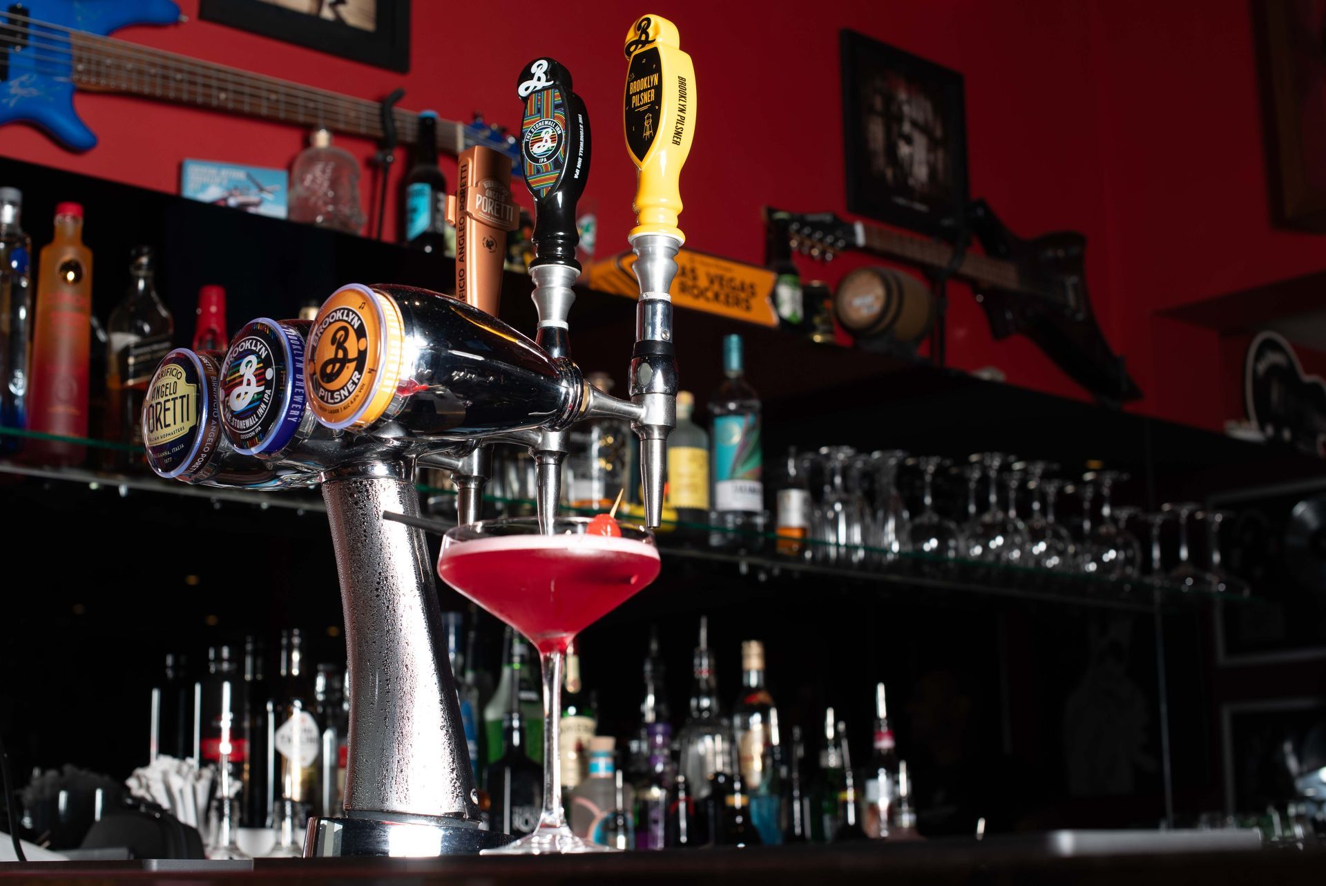 A bar with beer taps pouring a pink drink. Shelves behind the bar hold various bottles and glasses, with guitars mounted on the red wall.