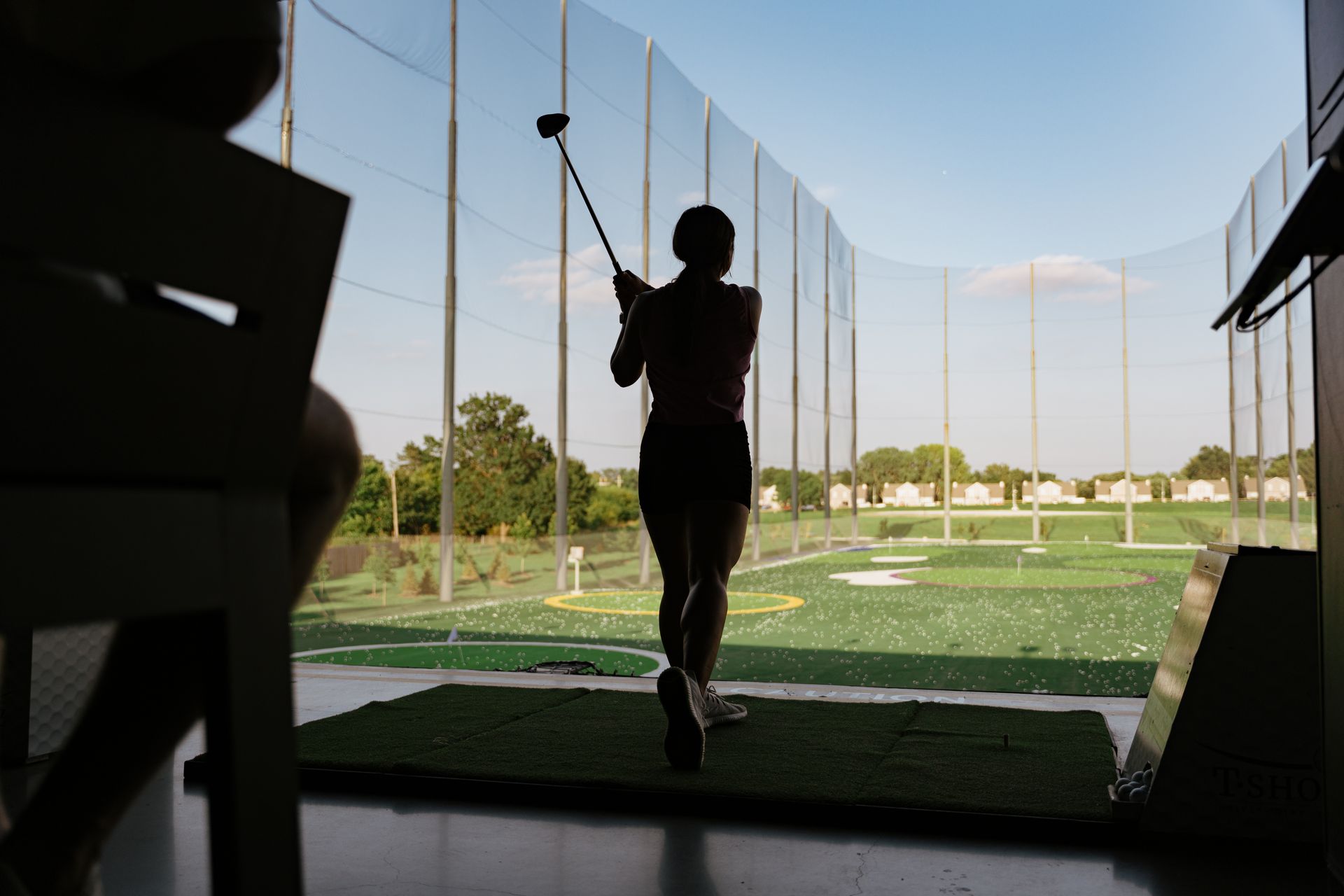 Silhouette of person swinging a golf club at a driving range, seen through a netting with a bright sky.