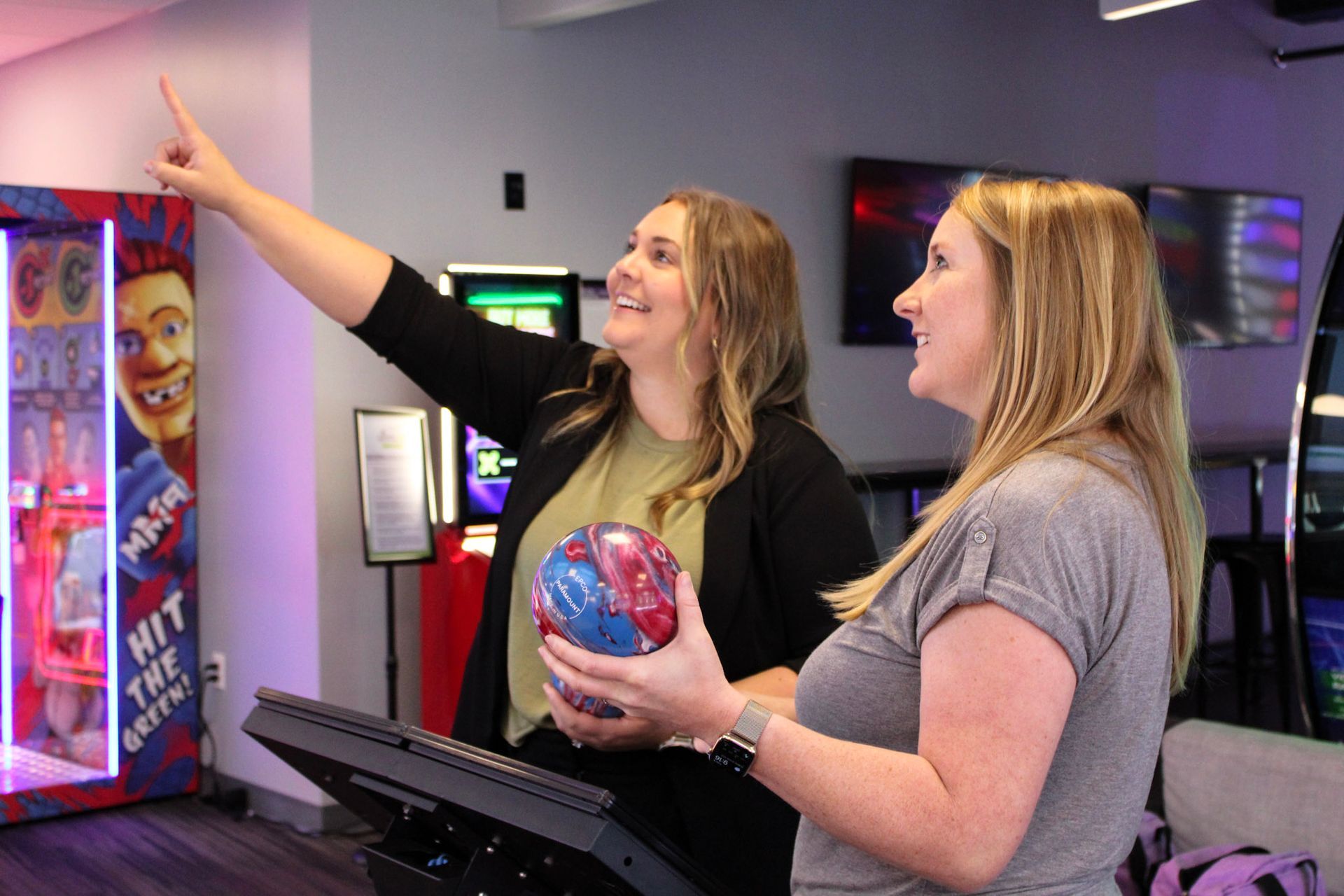 Two women in a bowling alley, one pointing up, holding a ball. Smiling faces, arcade in the background.