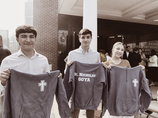 Three people holding dark shirts outside a building, with a cross emblem and one shirt reading “St. Richards Gutta.”