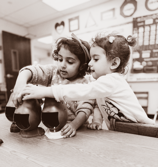 Two children sit on the floor, one helping the other adjust a shoe in a classroom-like room.