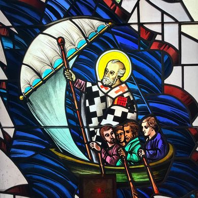 Stained-glass scene of a haloed saint in a boat with several figures amid blue waves and a sail