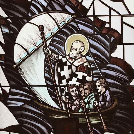 Stained glass of a saint in a boat, haloed and holding a staff, with figures below on blue waves.