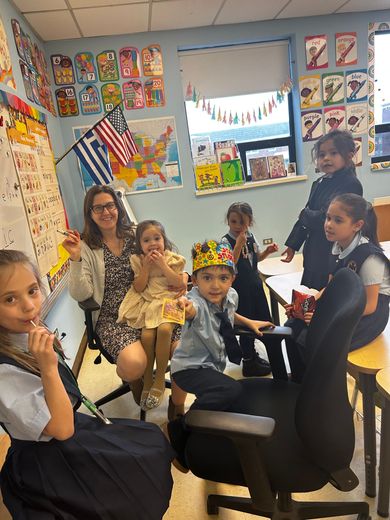 Teacher and children posing in a decorated classroom, with one child wearing a gold crown and others seated nearby.