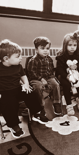 Three children sitting on a classroom rug, one holding a doll, in a black-and-white photo.