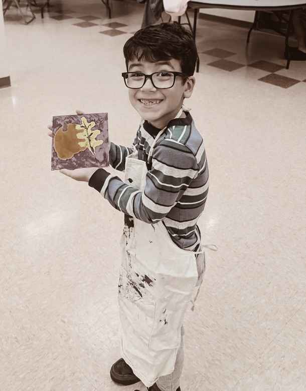 Smiling child in glasses holding a colorful artwork in a bright room, wearing striped shirt and paint-splattered apron