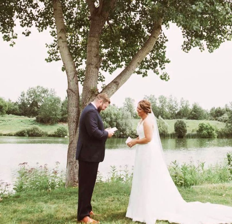 A bride and groom are standing under a tree near a lake