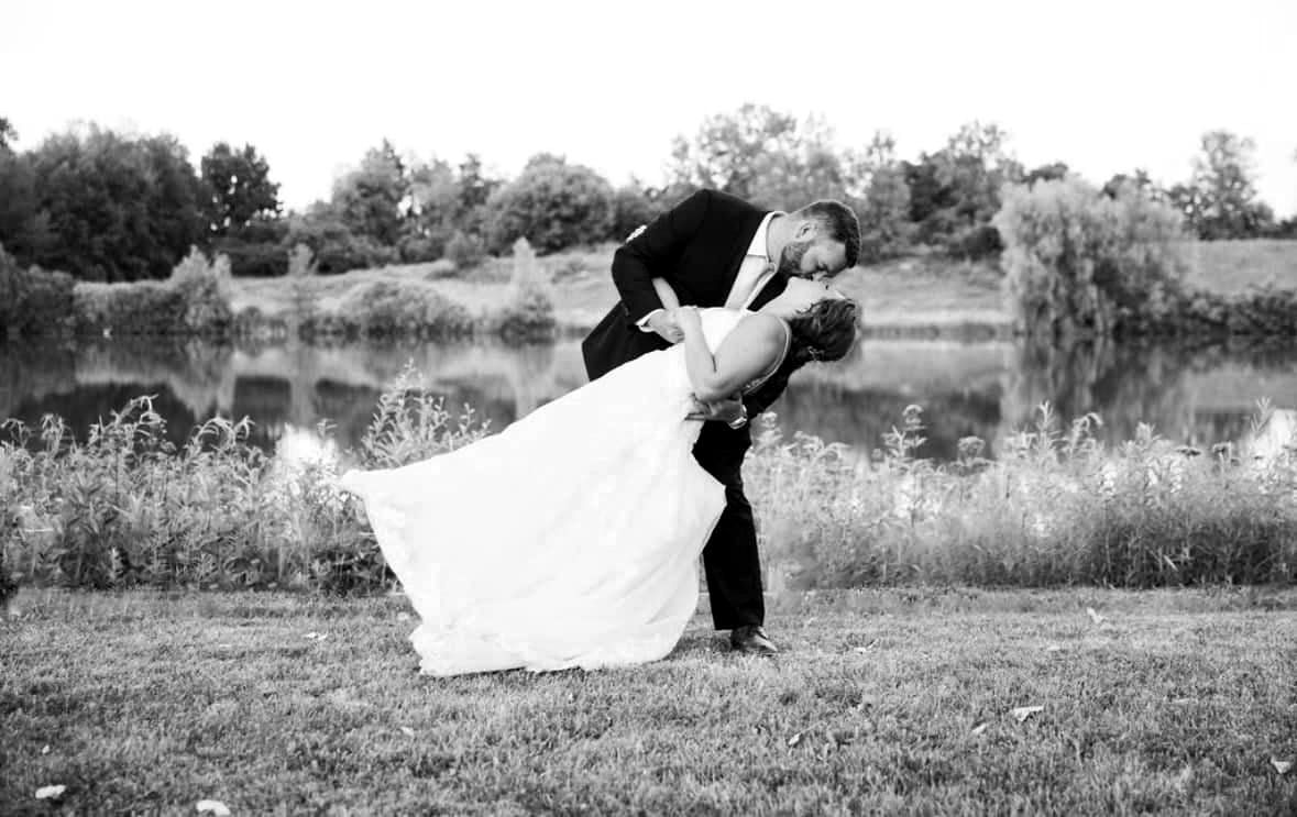 A bride and groom are kissing in front of a lake in a black and white photo.