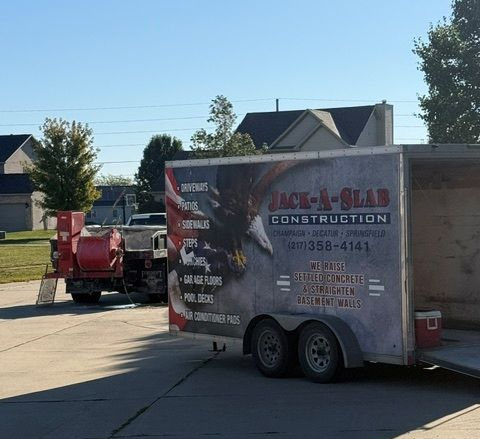 Utility trailer with Jack-A-Slab Construction branding parked in a suburban driveway beside a red machine