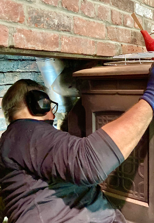 Brick chimney on a snow-covered roof, with fallen bricks. Background: a red brick building and trees.