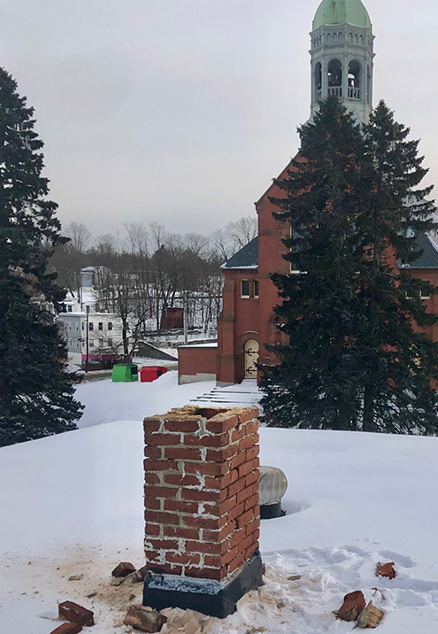 Person on a ladder painting a brick chimney against a blue sky. Yellow house visible.