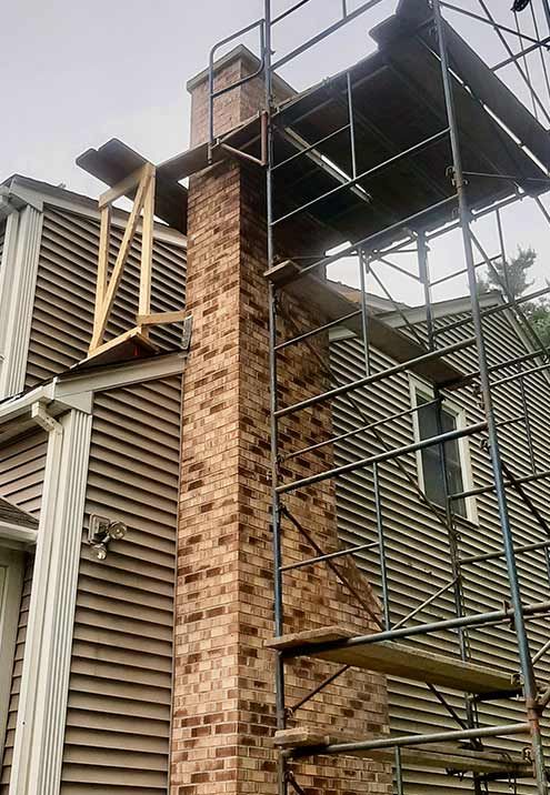 Brick chimney with scaffolding, attached to a brown-sided house. Construction work is in progress.