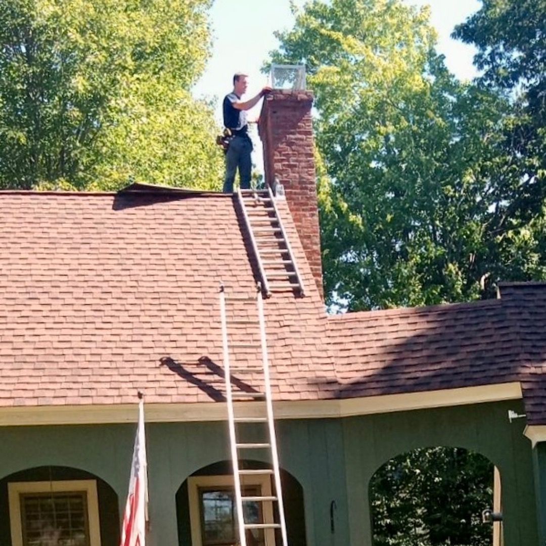 Man on a roof near a chimney, standing on a ladder. Green house, brown roof, trees.