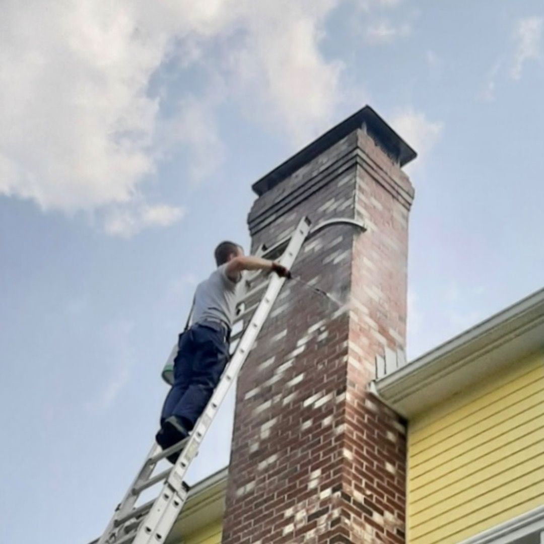 Person on a ladder painting a brick chimney against a partly cloudy sky. The house is yellow.