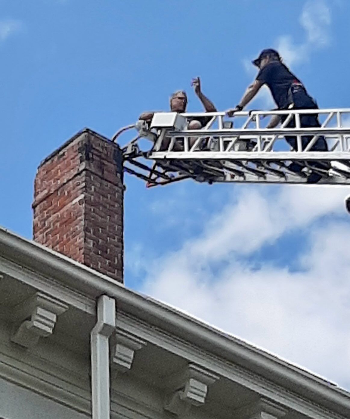Firefighters on a ladder near a brick chimney. One is reaching up; blue sky.