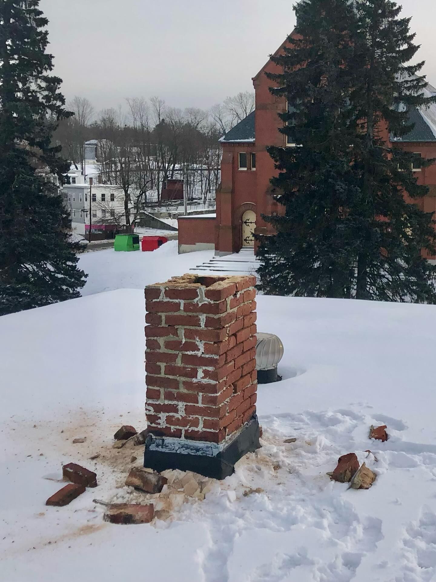 Brick chimney on a snow-covered roof, with fallen bricks. Red brick building in background.
