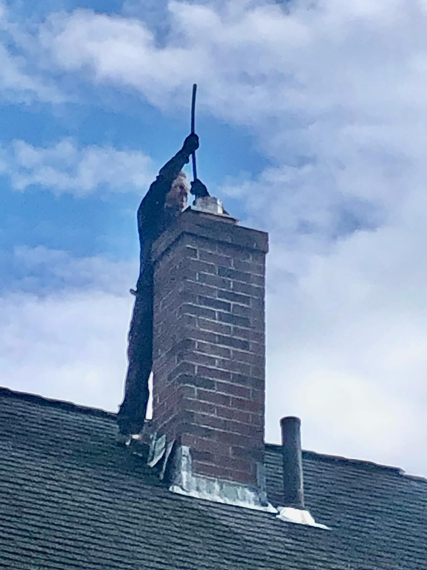 Chimney sweep on a rooftop cleans a brick chimney with a long rod against a cloudy sky.