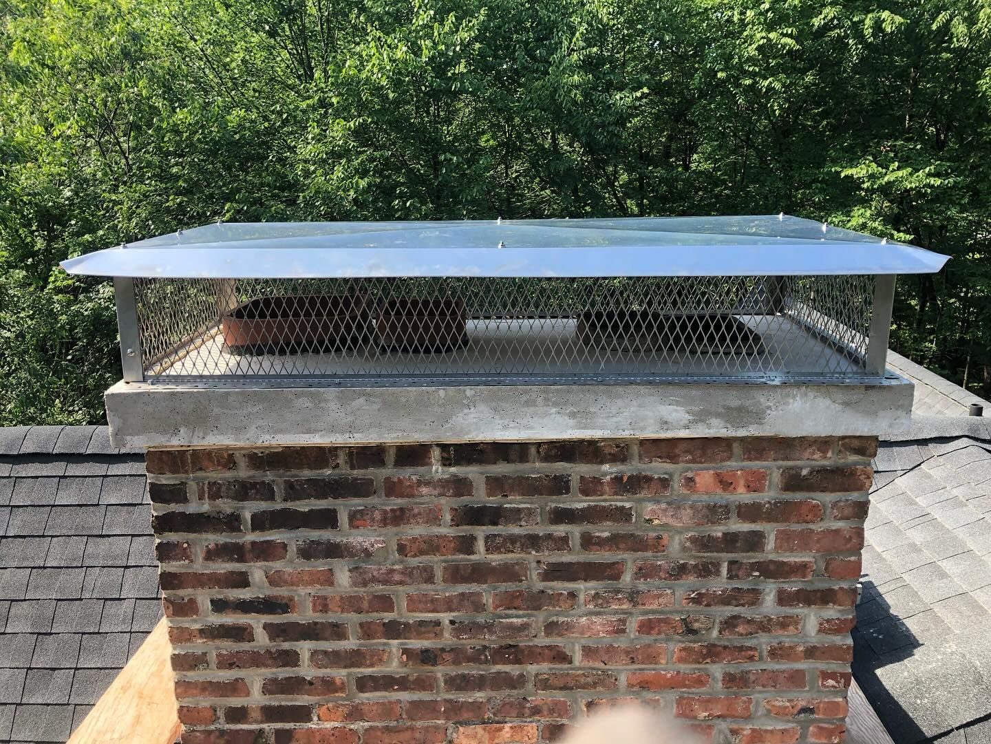 Brick chimney with concrete cap and metal chimney cap on a shingled roof, trees in the background.