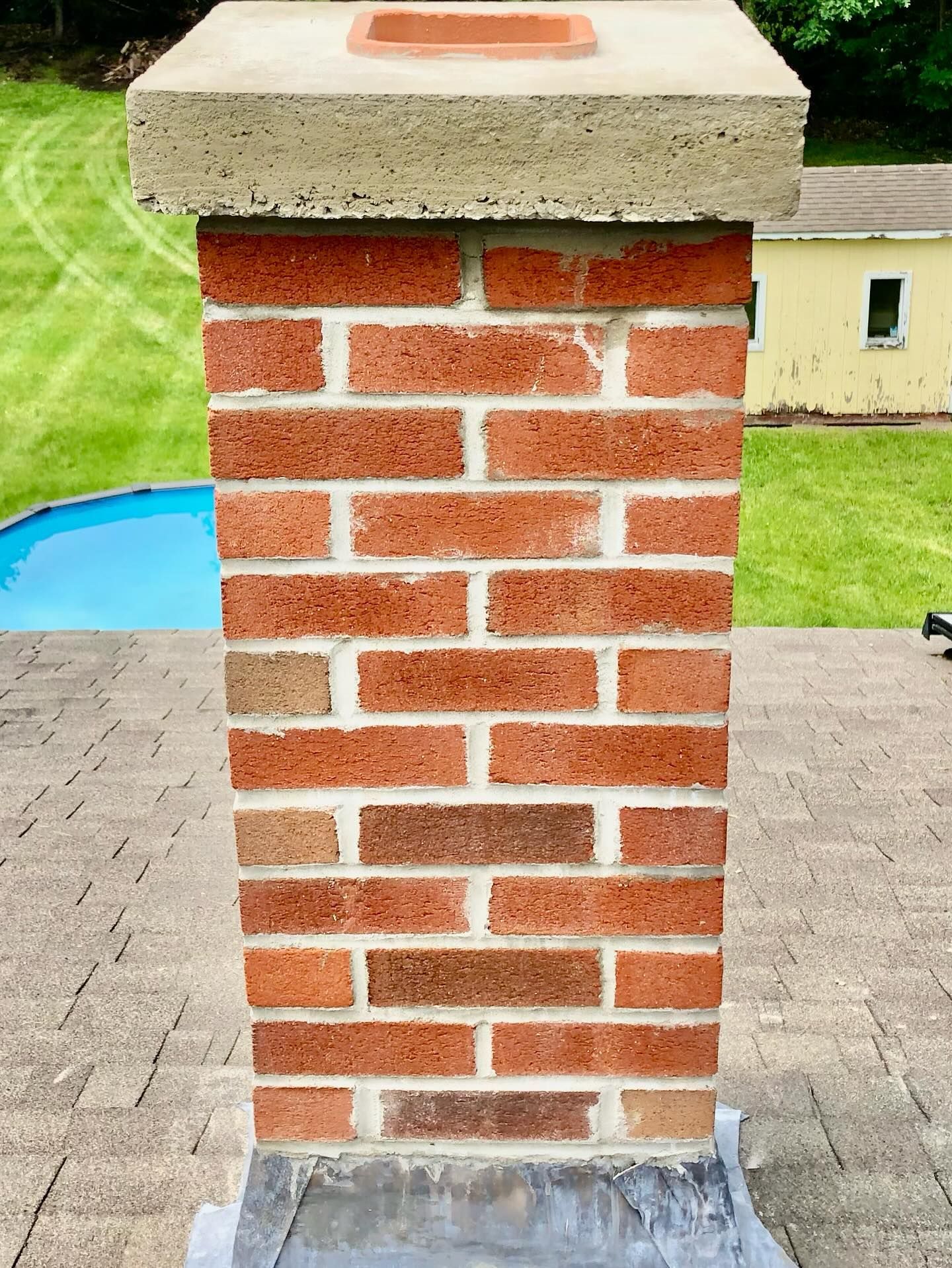 Brick chimney with concrete cap on a roof, blue pool and yard in the background.