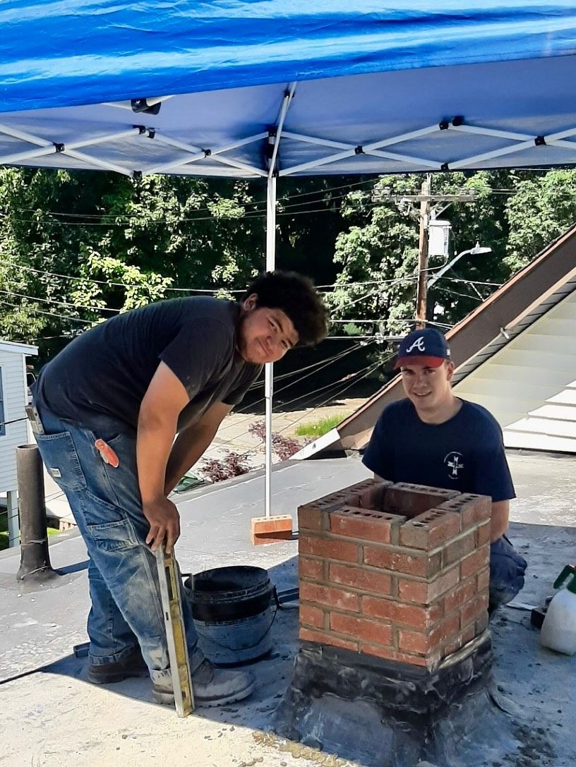 Two people building a brick chimney on a rooftop under a blue tarp.