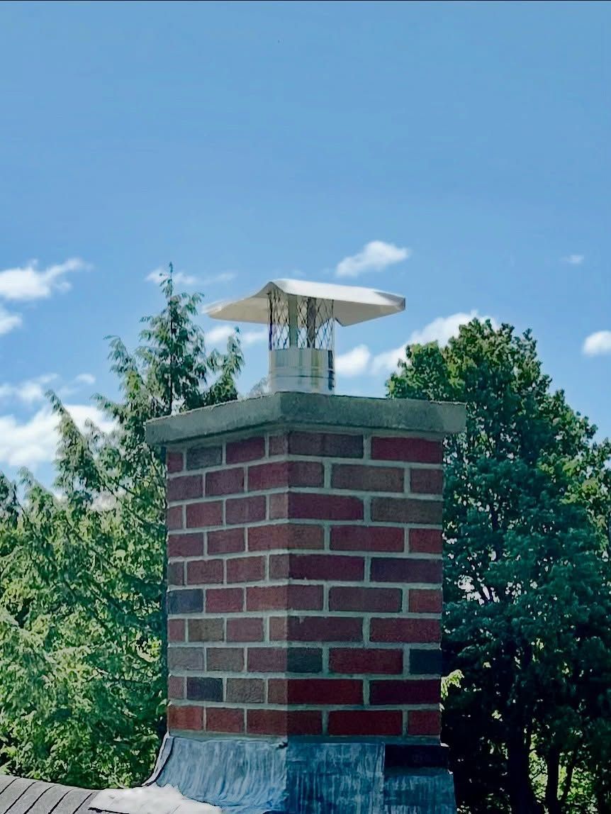 Brick chimney with metal cap, on a rooftop with trees and blue sky with clouds.