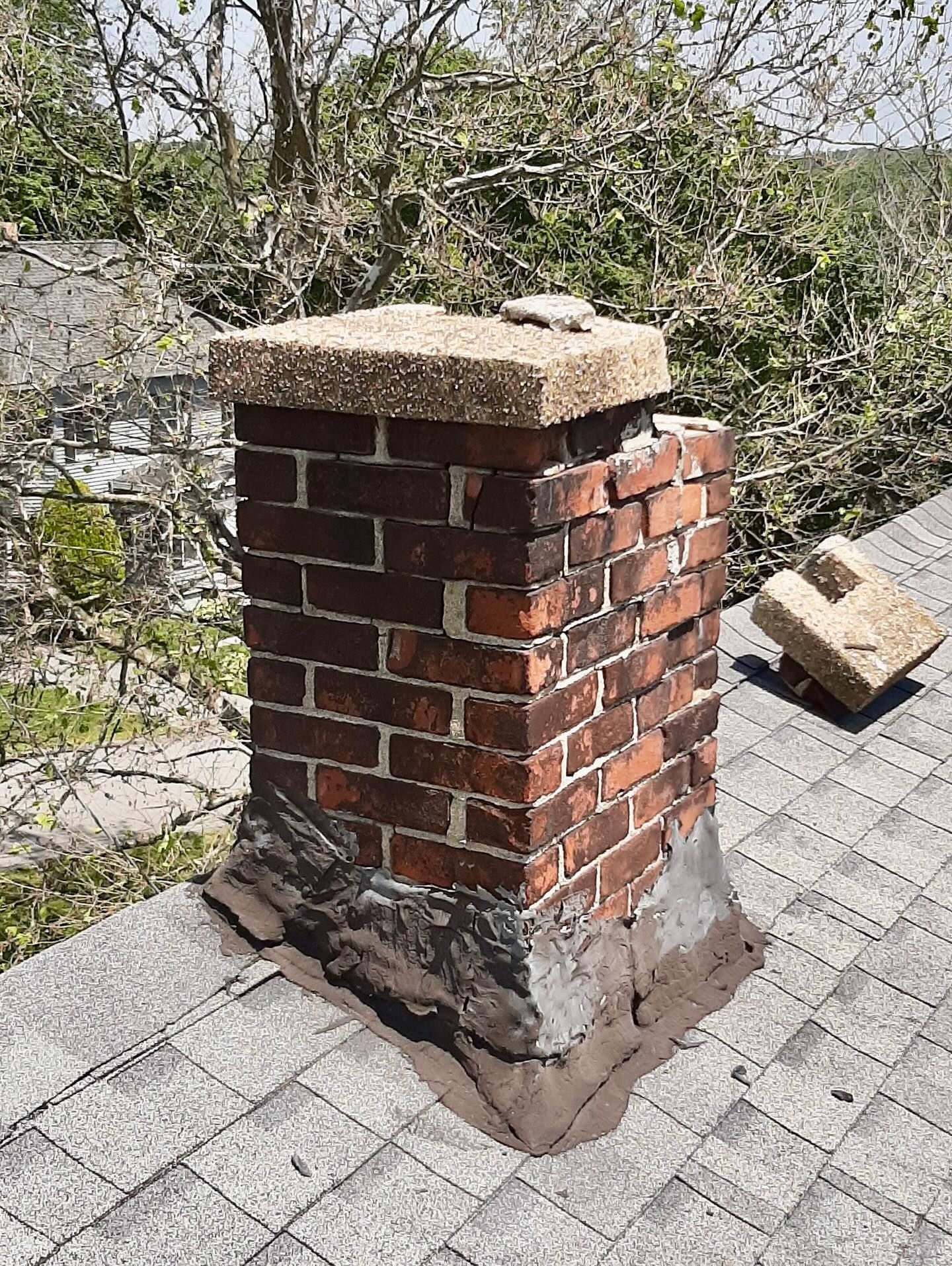 Brick chimney on a gray shingled roof, with mortar damage and a detached cap stone.