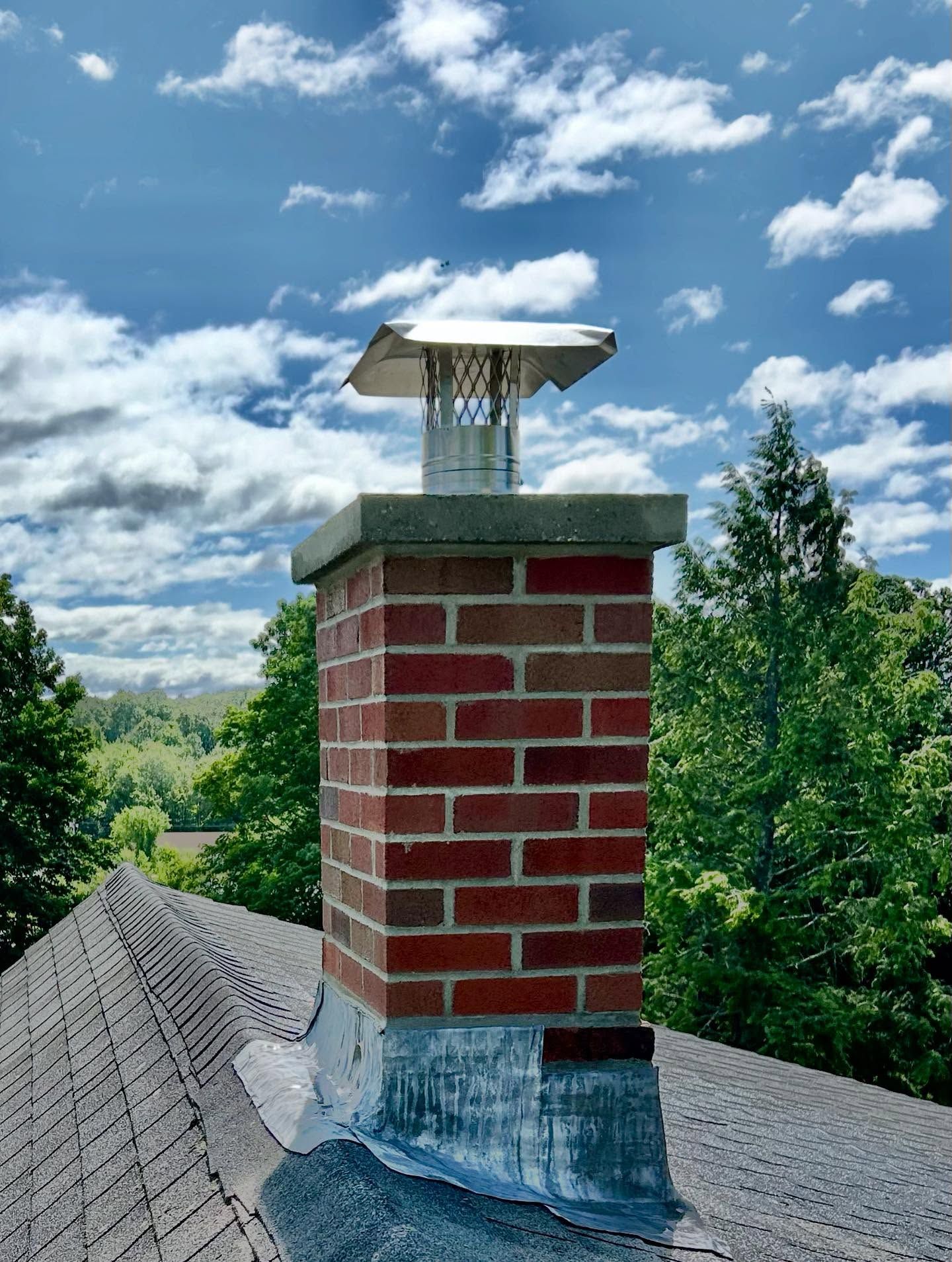 Brick chimney on a roof with a metal cap, against a cloudy blue sky.