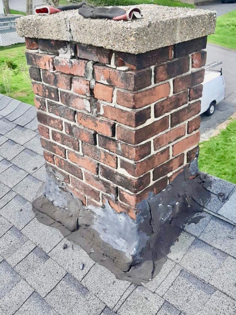 Brick chimney on a gray shingle roof, showing cracks and damaged flashing.