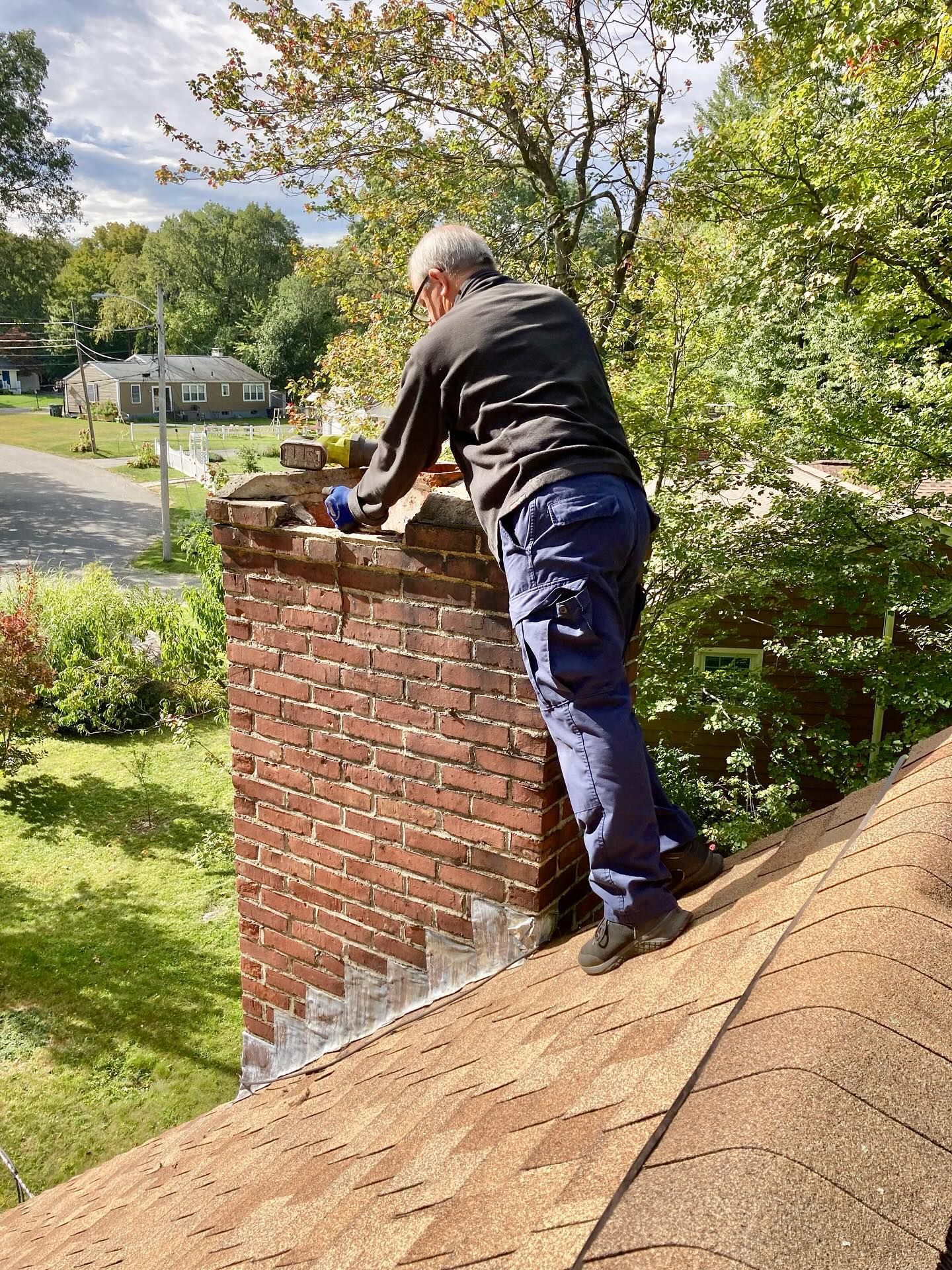 Person repairs a brick chimney on a rooftop. They wear blue pants and a black shirt. Green trees and sky in the background.