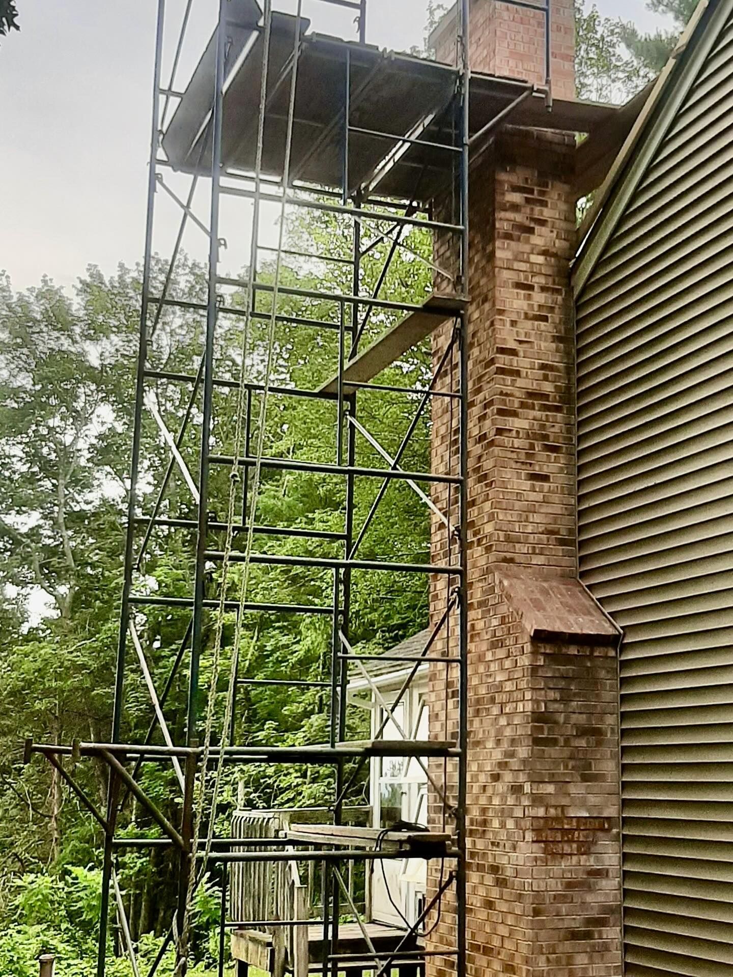Scaffolding next to a brick chimney of a house for repairs; green trees are in the background.