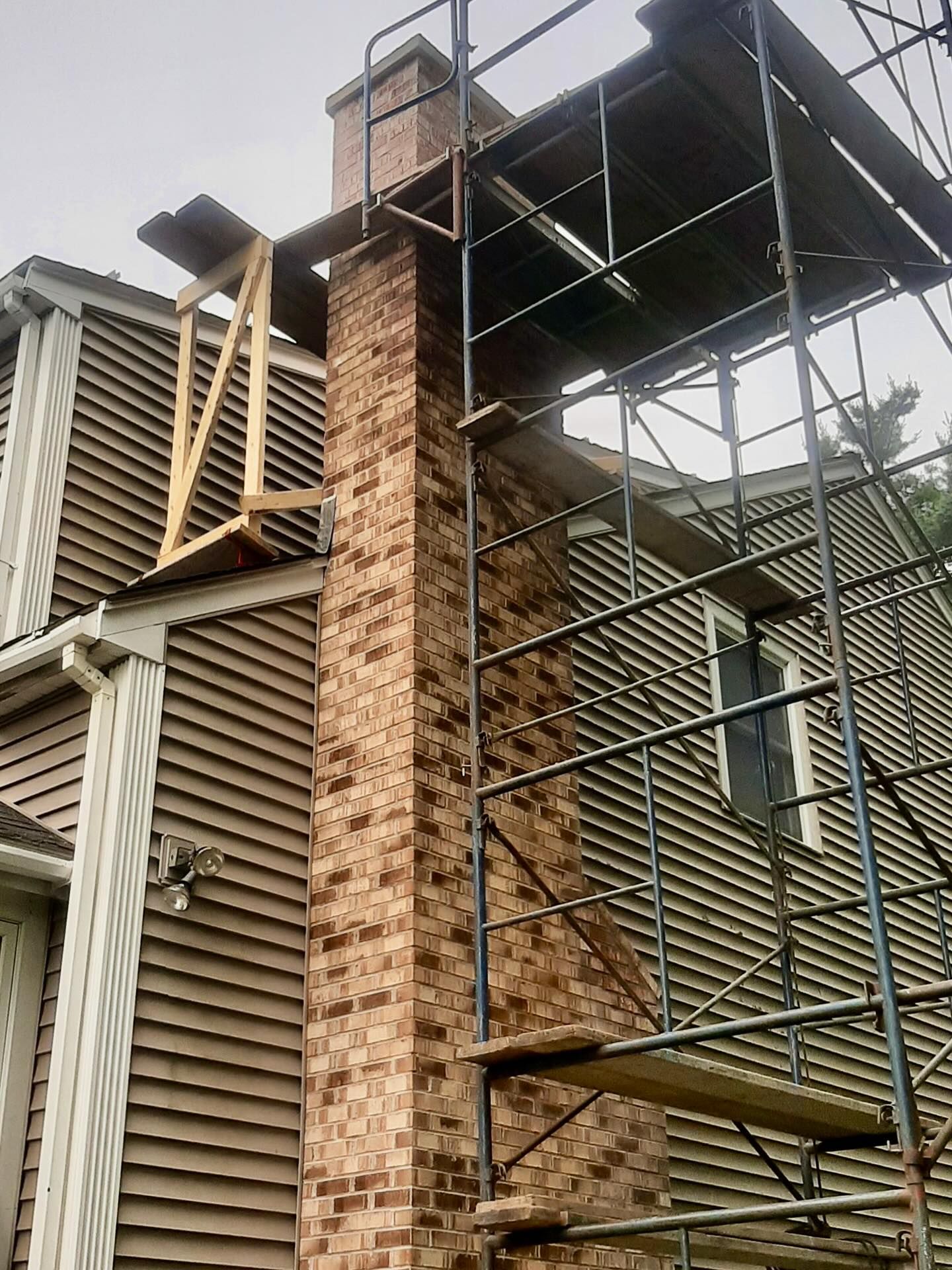 Brick chimney with scaffolding next to a brown-sided house.