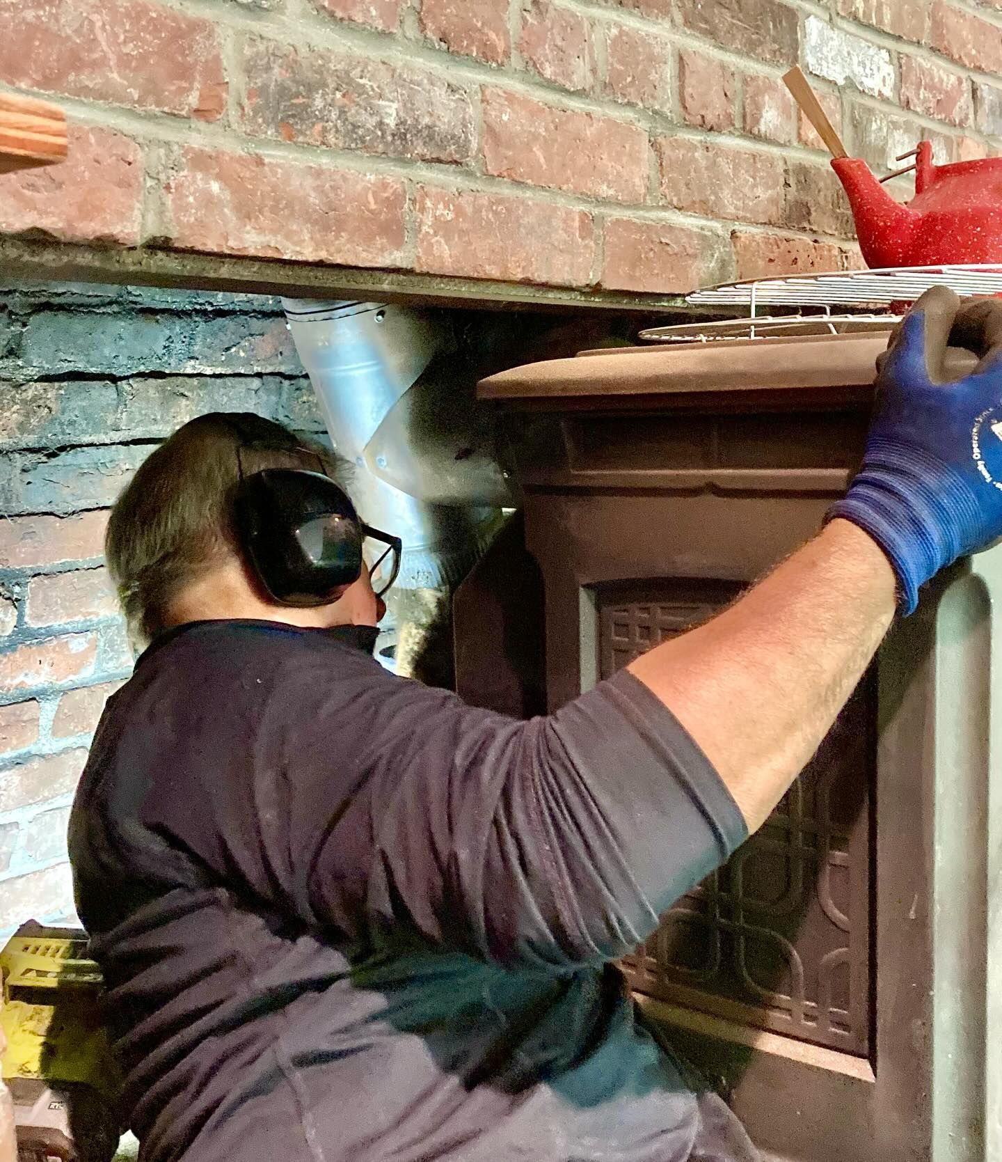 Man wearing ear protection and gloves working on a stove beneath a brick wall.