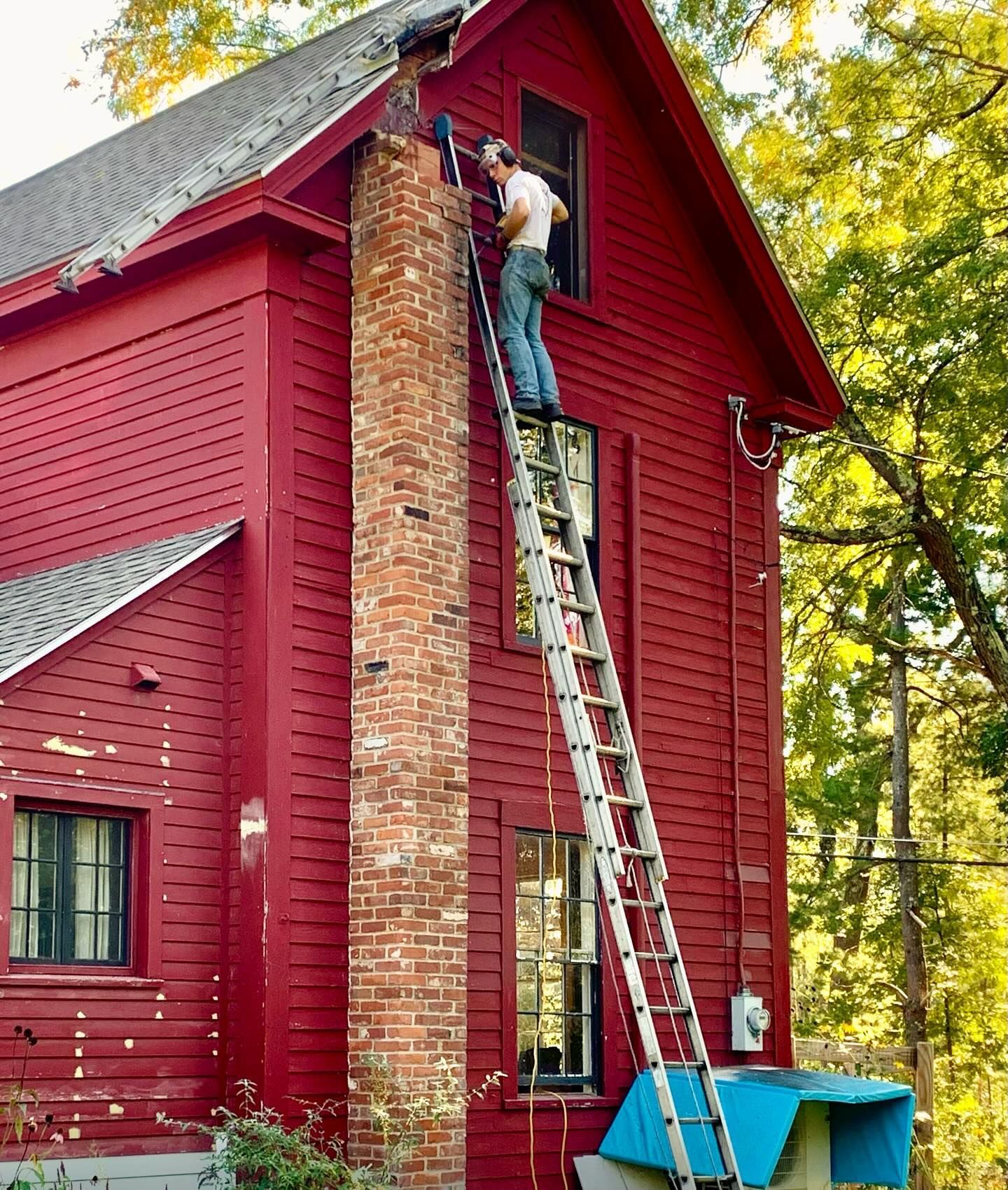 Man on a tall ladder repairing a brick chimney on a red house.
