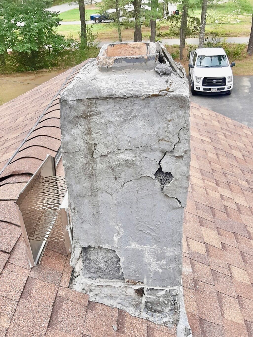 Damaged chimney on a brown shingle roof, with a white truck parked in the background.