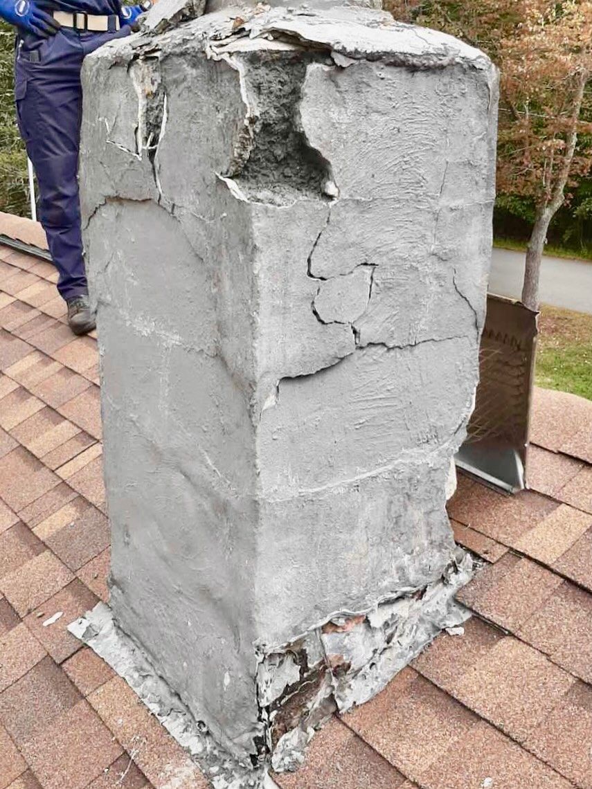 Damaged chimney with crumbling gray coating on a brown shingle roof, person in blue pants in background.