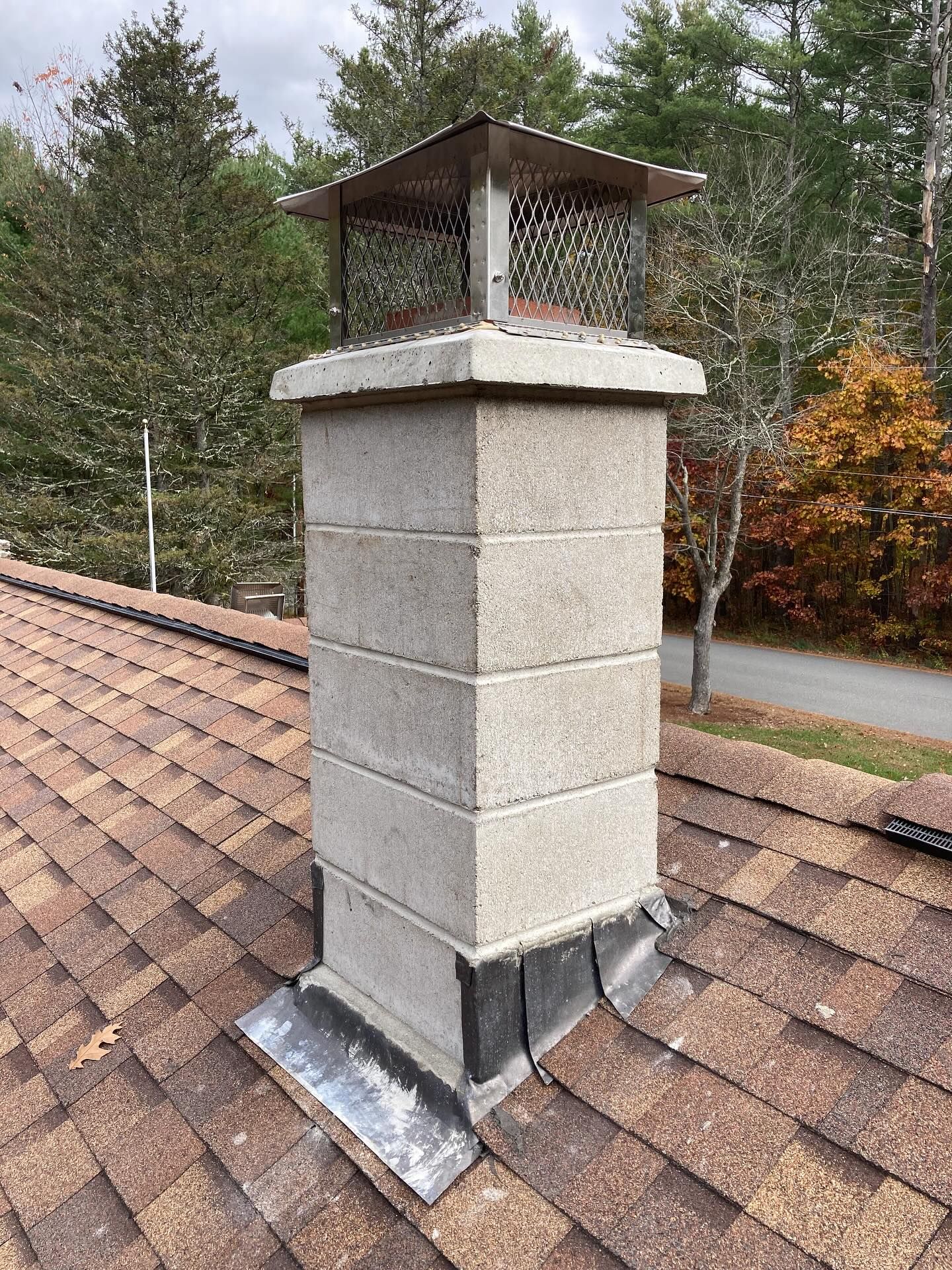 Gray block chimney with metal cap on a brown shingle roof, surrounded by trees.